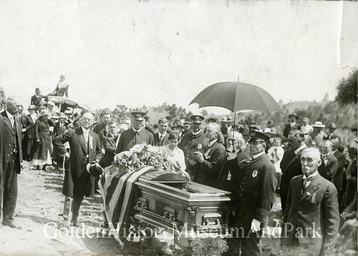 A group of people gathered around a coffin, one holding an umbrella for shade.