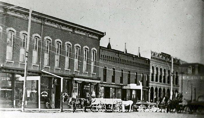 Black and white photo showing the west side of the 1200 block of Washington Avenue. The Opera House, Harrison block, and Everett block are included. The street is unpaved and a horse and wagon wait in from of the Opera House.
