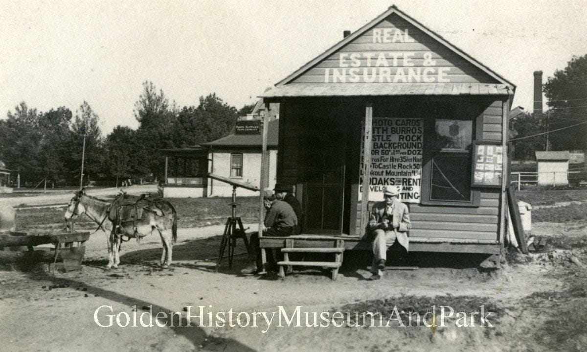 Two men sit on the porch of a small wooden building with Real Estate & Insurance painted over door. Burro tied near by.