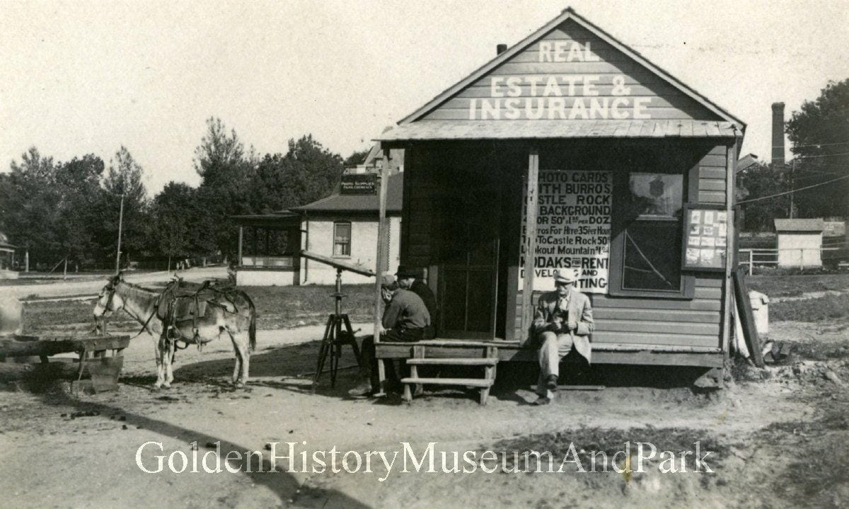 Two men sit on the porch of a small wooden building with Real Estate & Insurance painted over door.  Burro tied near by.