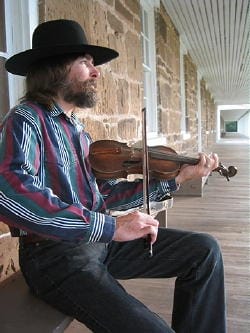 Bearded man in a cowboy hat sits on a bench in front of a stone building, playing a violin.