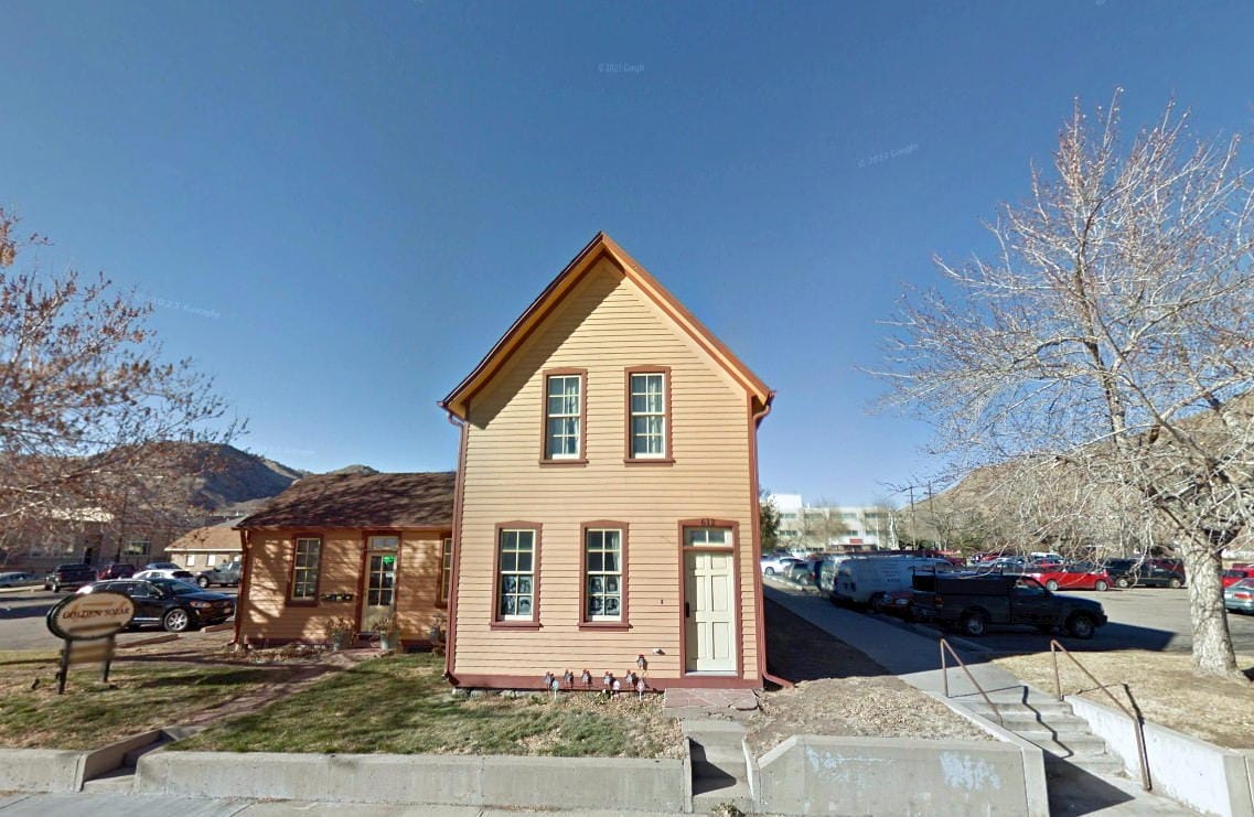 2 story Victorian house with a 1 story el, surrounded on three sides by parking lots with CoorsTek in the background