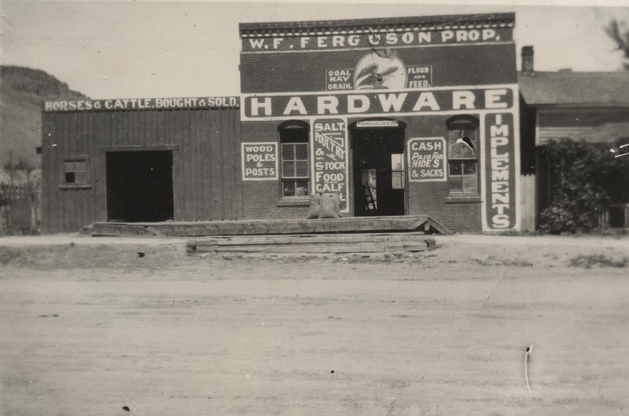 Wooden store building with large sign saying HARDWARE, smaller signs for horse & cattle bought & sold, coal, hay, grain
