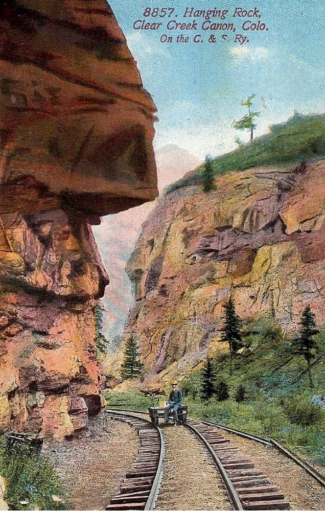 Big rock jutting from the canyon wall hangs over a man sitting on a handcart on narrow gauge tracks.