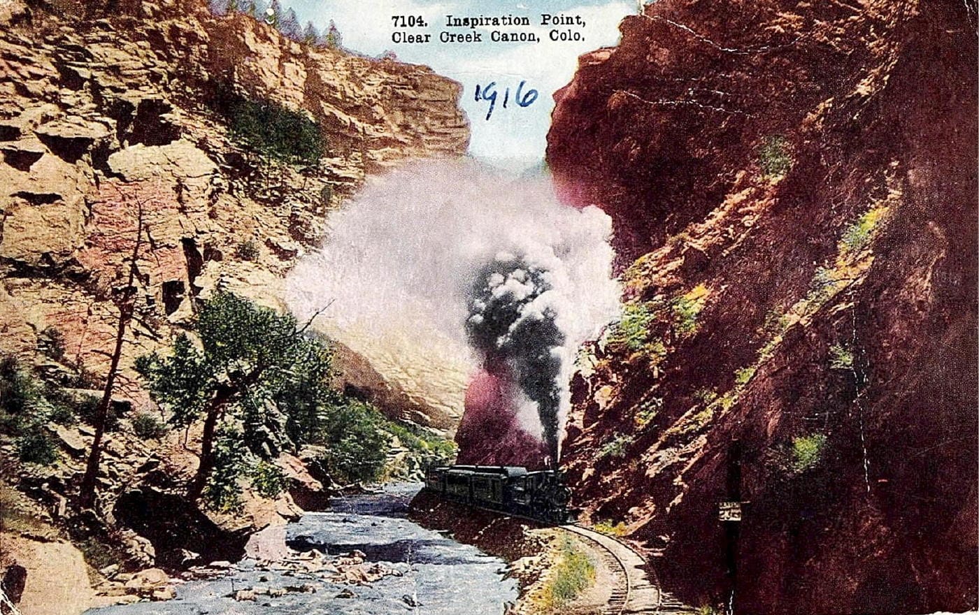 Steam locomotive coming down the tracks toward photographer.  Red canyon walls and big black plume of smoke from the train.