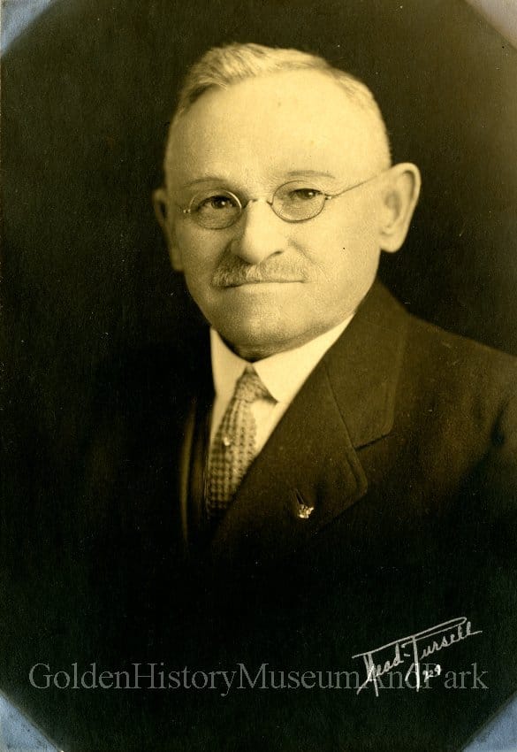 man with mustache, gray hair, and wire-rimmed glasses wearing a suit with tie tac and lapel pin