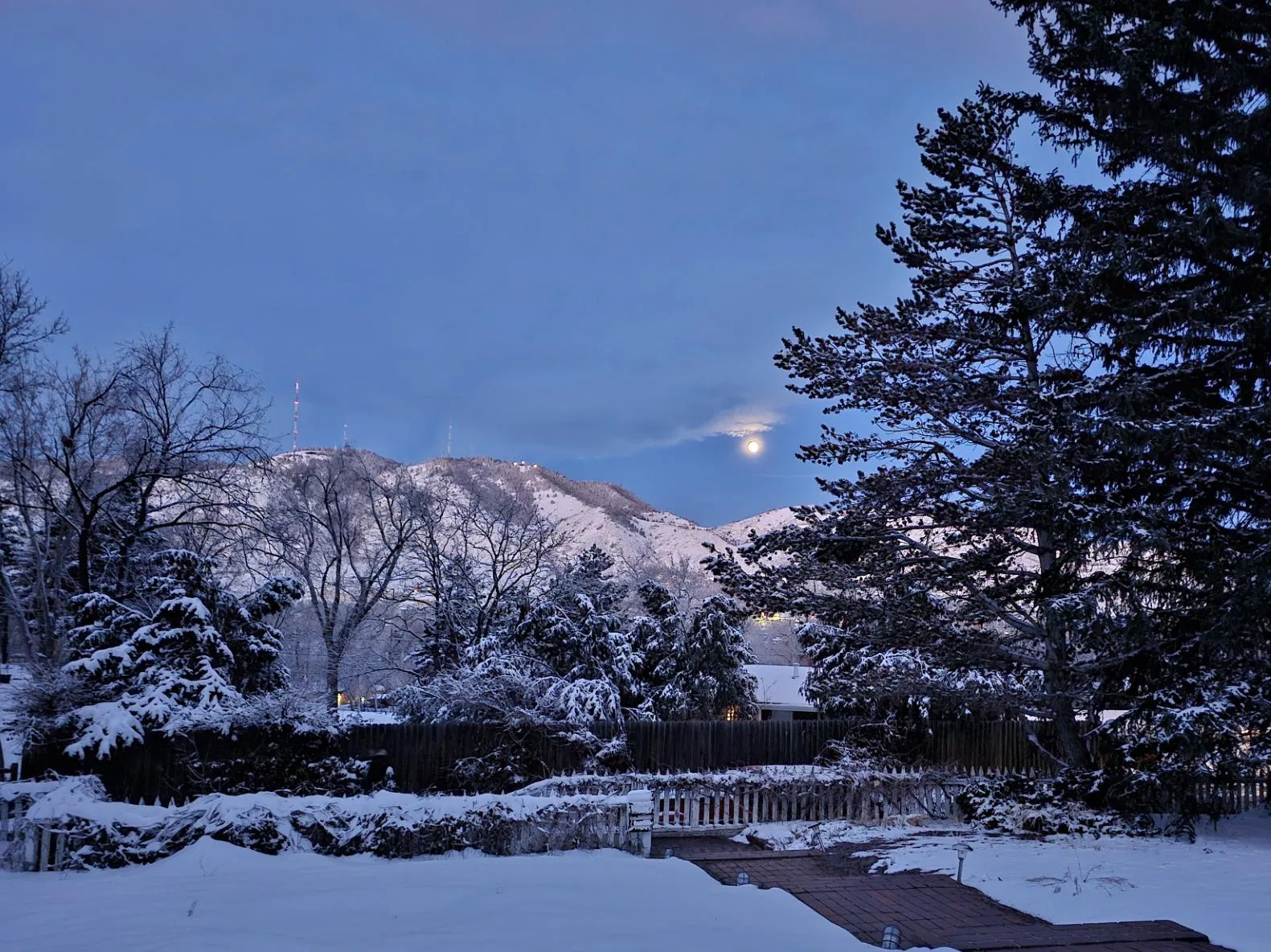 Full moon setting behind snow-covered foothills.  Brick sidewalk and large evergreens in foreground.