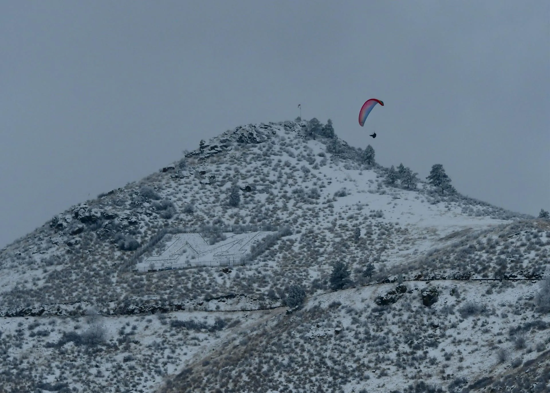 Hang glider with a brightly colored sail drifts near the peak of Mount Zion on a dark, snowy day.