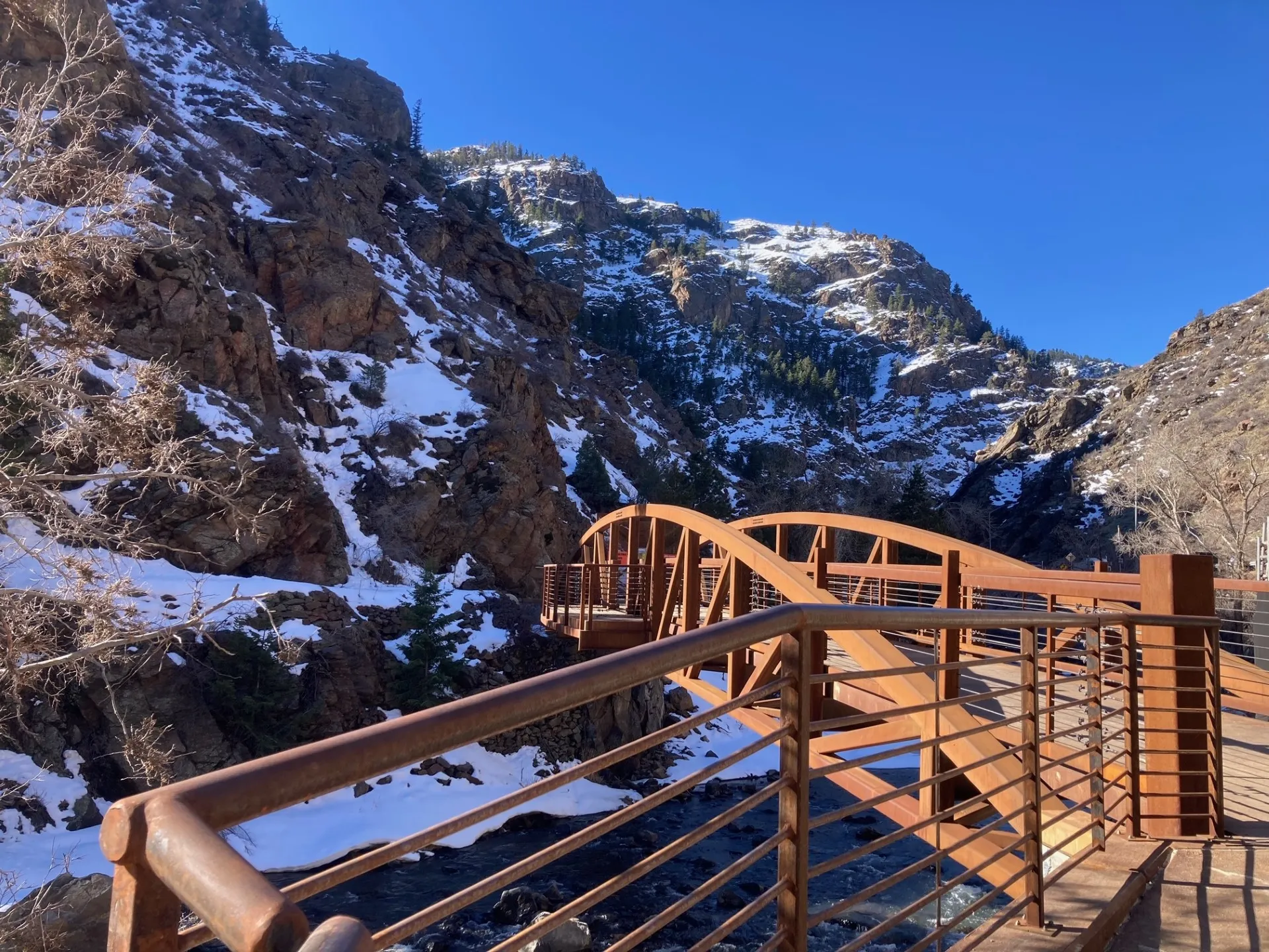 Brilliant blue sky over a canyon with snow clinging to the walls.  Brown footbridge goes over Clear Creek.