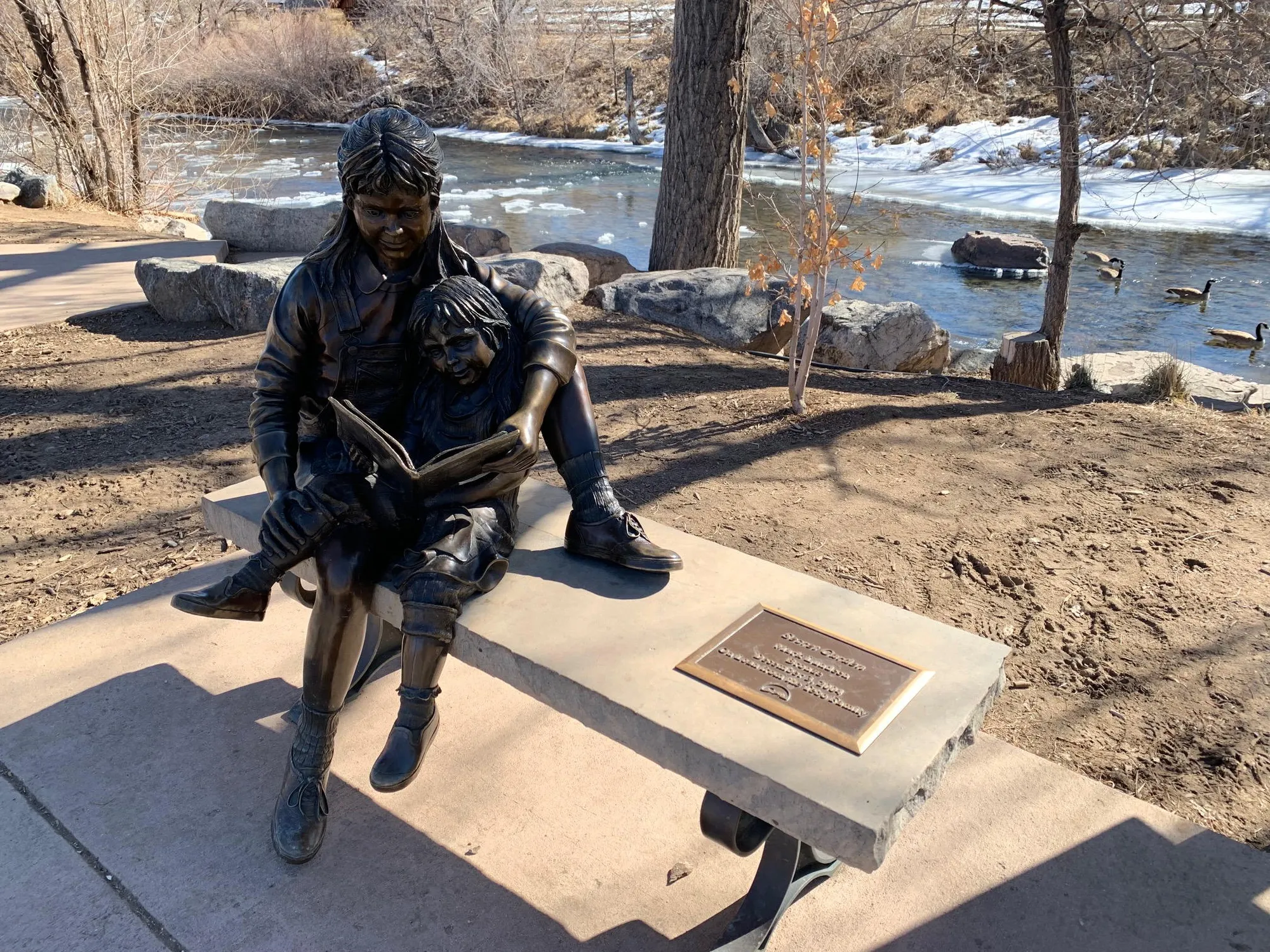 Bronze statue of children reading between the Golden Library & Clear Creek.  Geese in Creek, ice along edge, & snow on bank.