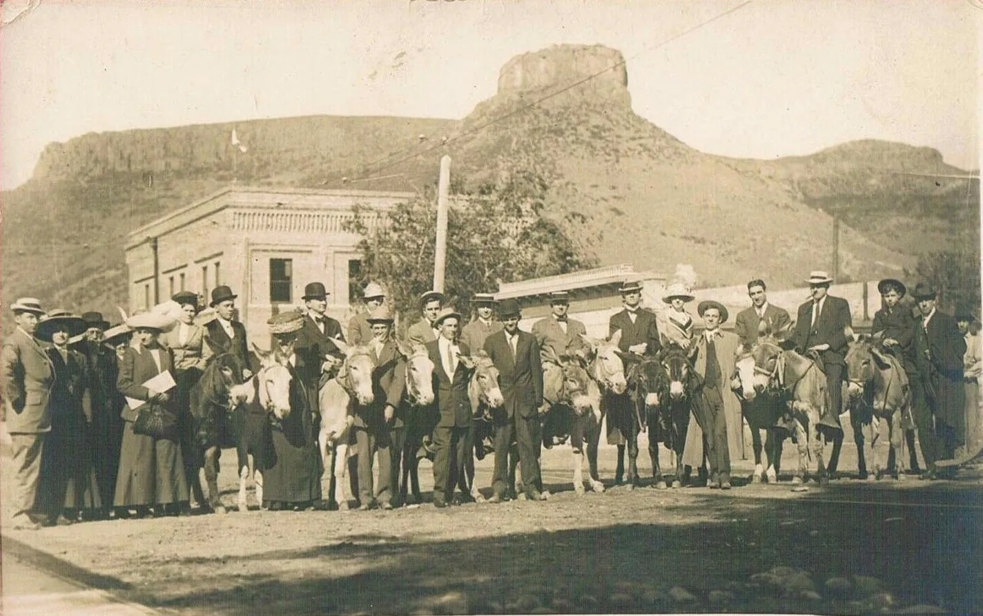 A wide group of circa-1910 tourists, about 11 seated on burros and the rest on foot.  Castle Rock in the background.