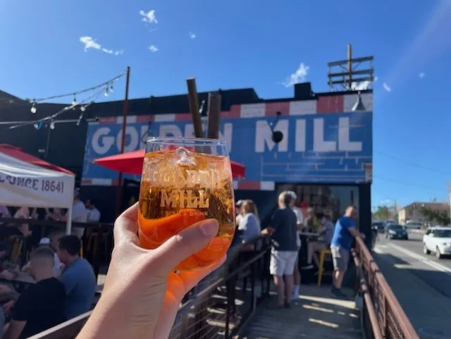 a hand holds aloft a Golden Mill glass, filled with pinkish liquid and ice.  The Golden Mill sign appears in the background.