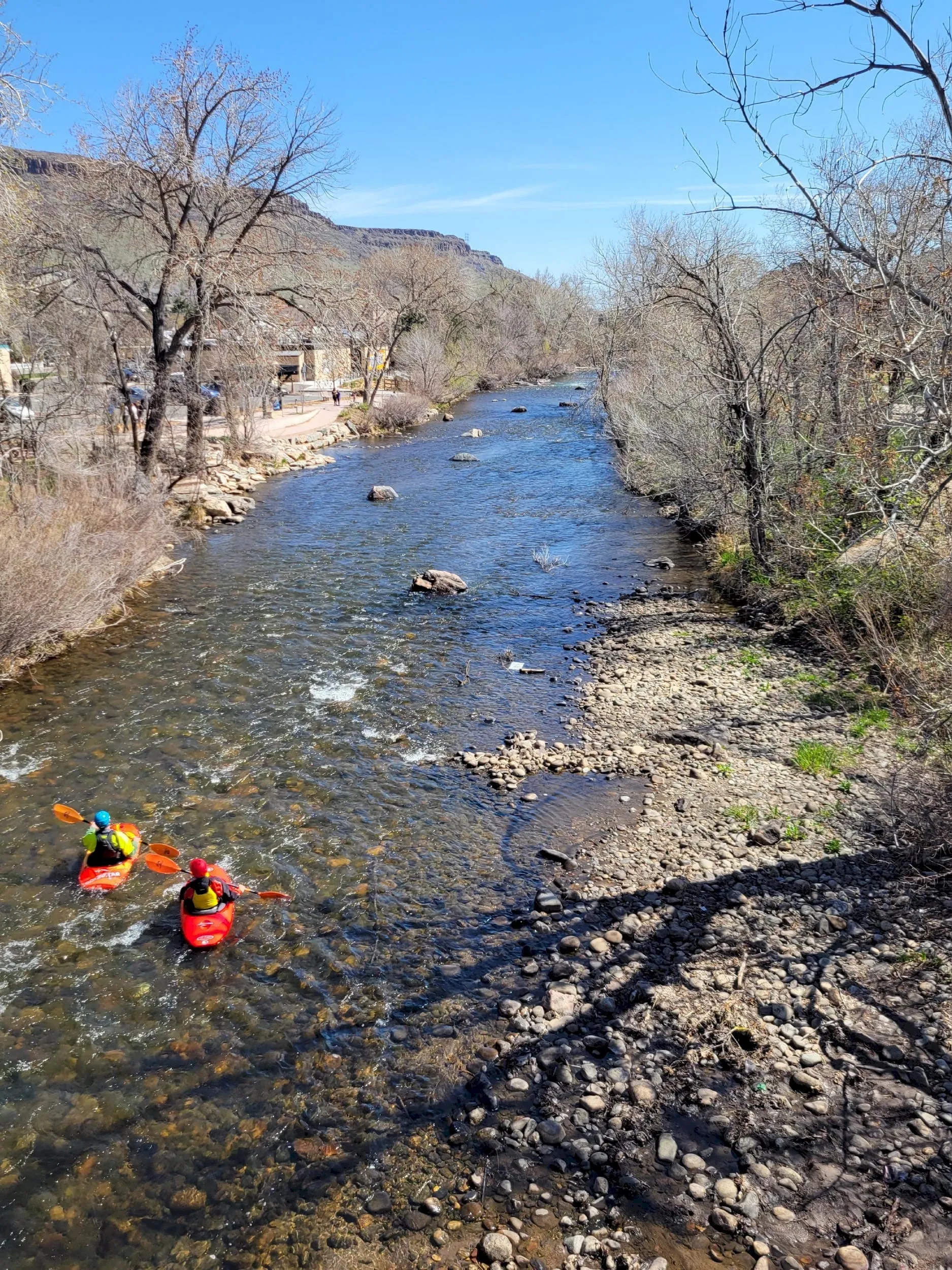 Looking down on a very shallow Clear Creek with two kayakers in red crafts.  Trees are bare but some grass is greening.