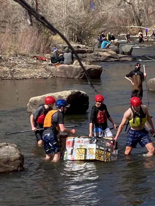 Young people wading in Clear Creek, standing around their cardboard boat. Spectators sitting along the Creek.