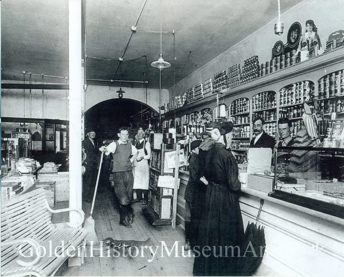 old-fashioned grocery store with merchandise behind the counter, two women patrons and five men, apparently employees