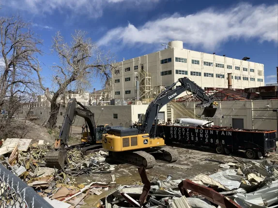 heavy equipment dropping construction materials into a large dumpster, the wreckage of a building, and some large, bare trees
