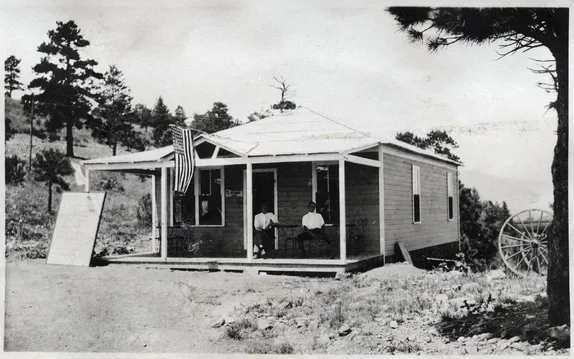 Two men sitting at tables on the porch of a small frame building.  Sign board leans against porch post.