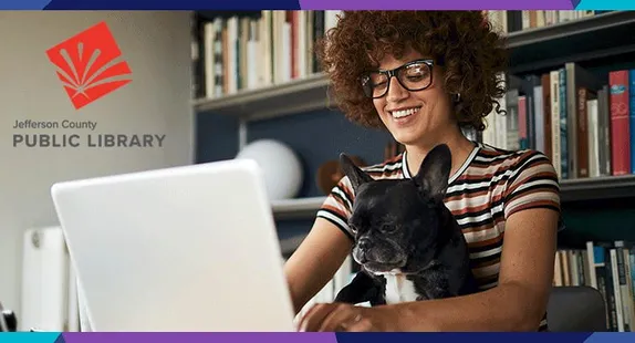 smiling woman with curly hair and glasses typing on laptop with dog under one arm, library shelves in back