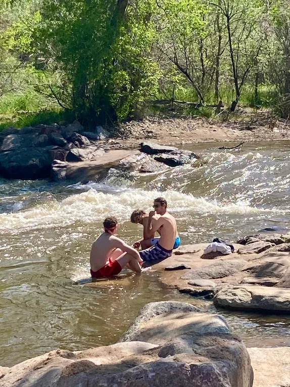 three young men in swimming trunks sitting in shallow creek waters