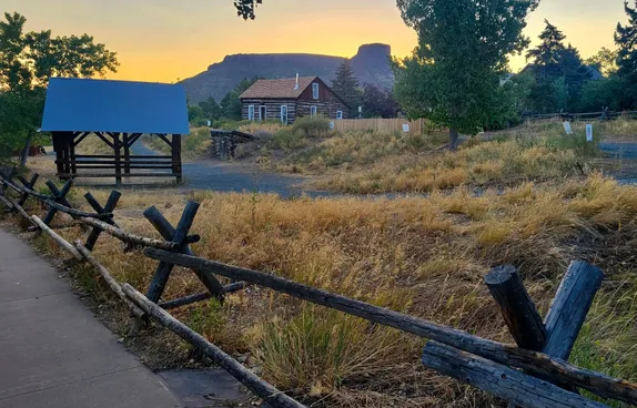 looking over the split rail fence at grass, a hay barn, a cabin, and castle rock in the background