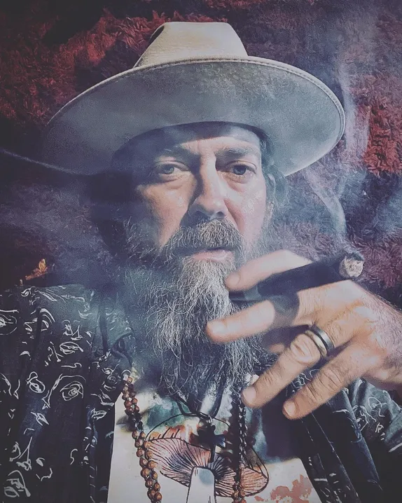bearded man in cowboy hat smoking cigar - wearing beads with mushroom picture on his shirt