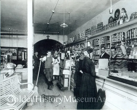 old-fashioned grocery store with merchandise behind the counter, two women patrons and five men, apparently employees