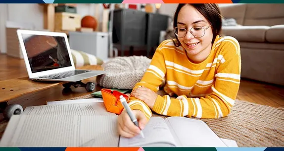 Young girl writng in a journal with a laptop computer nearby. 